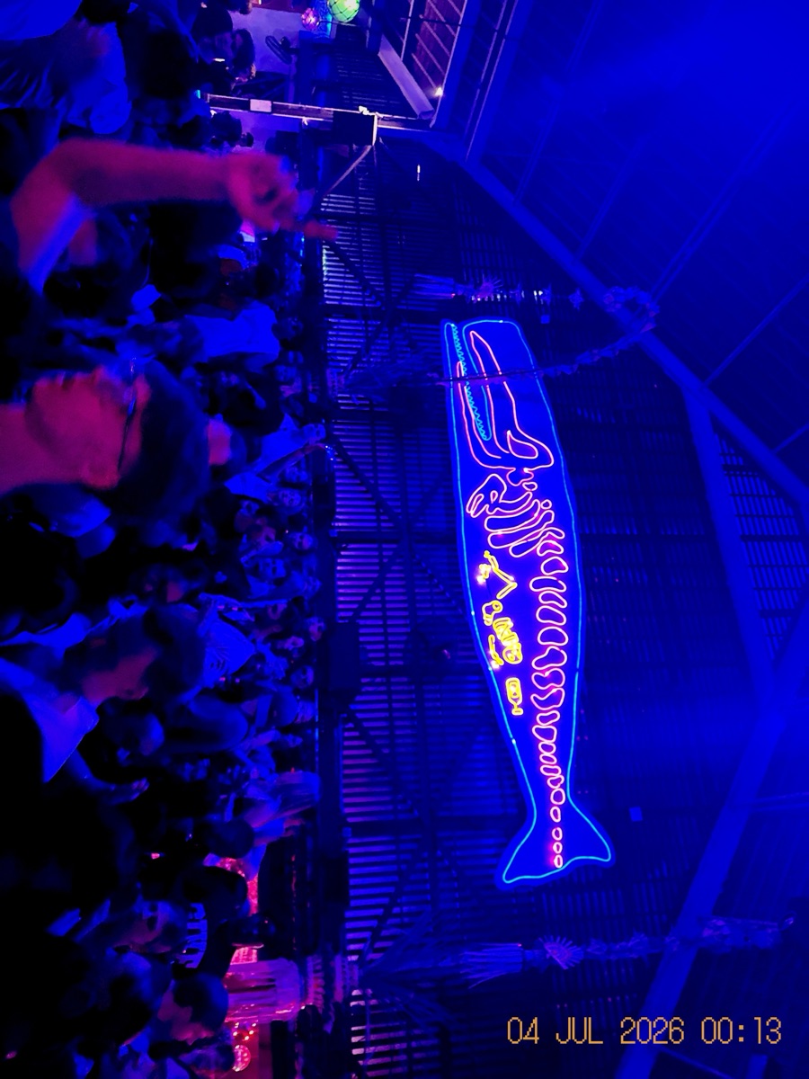 A neon fish sign above a crowd at night, shot on the Chrome film stock: intense saturated blue, crisp, no grain.